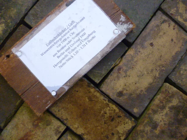 Metal plaque mounted on a wooden frame lying on damp paving stones; contains German text.