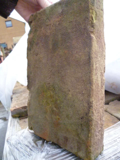 Moss-covered, weathered rectangular brick held upright by a hand, with a construction site backdrop and plastic sheeting nearby.