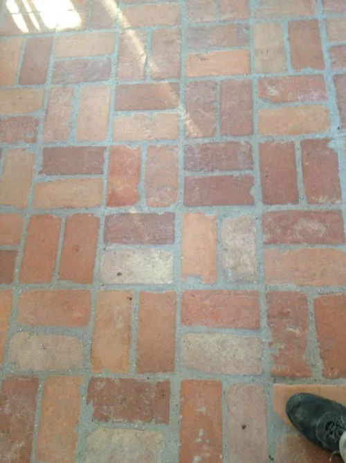 Brick floor with red and tan rectangular bricks in a staggered pattern; bottom-right corner shows part of a dark shoe.