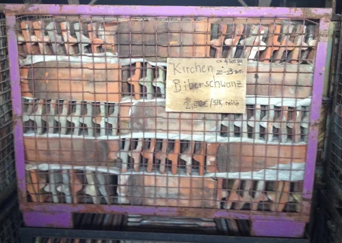 Wire-framed, purple-metal crate filled with assorted terracotta bricks and clay tiles, with a handwritten sign advertising kitchen bricks at €2.50 per piece.
