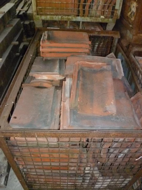 Stack of dusty red clay roof tiles in a rusted metal cage, stored in a dim, cluttered basement-like area