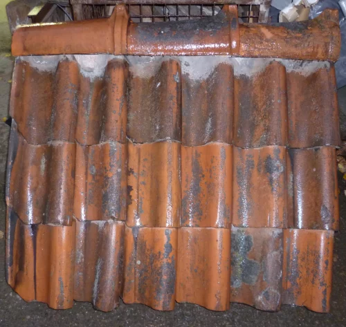Rows of curved orange terracotta roof tiles stacked in a workshop or storage area, showing weathering and rust-like stains.